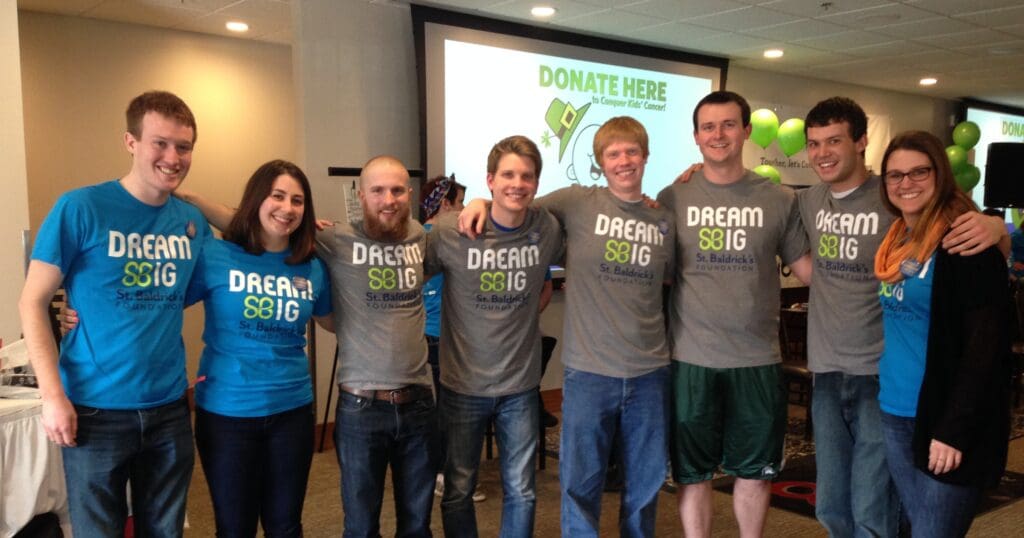 Eight smiling people wearing "Dream Big" shirts pose together indoors. A banner behind them reads "Donate Here," suggesting a fundraising event.