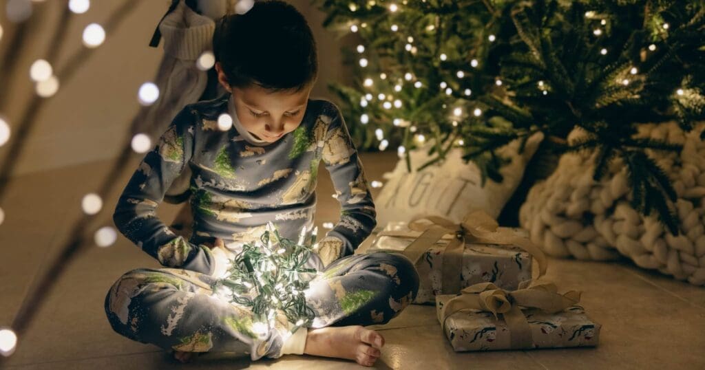 A child in pajamas sits on the floor, holding glowing Christmas lights. They are surrounded by wrapped presents and a lit Christmas tree, creating a warm, festive ambiance.