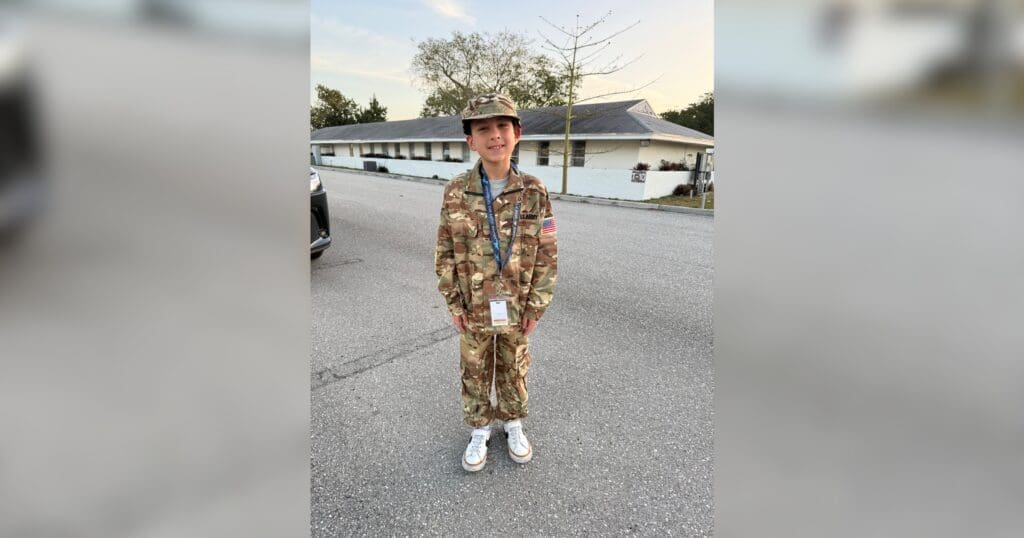 Young person wearing a camouflage uniform stands outdoors on a street, smiling. The mood is cheerful. The building and trees are in the background.