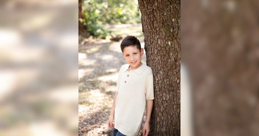 Young boy with dark hair leans against a large tree in a sunlit forest, smiling softly. He wears a beige polo shirt, conveying a serene, playful mood.