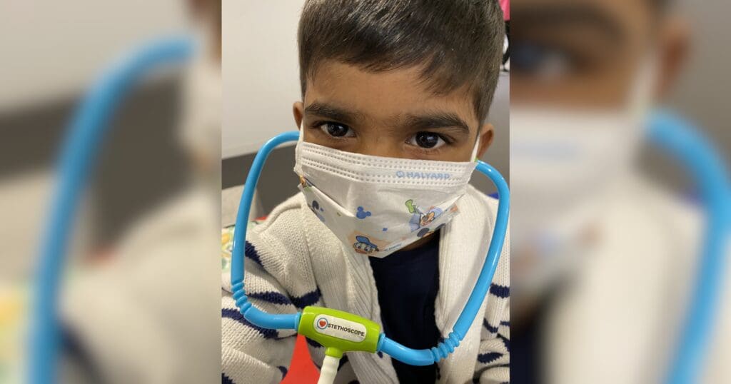 Young child wearing a face mask and a blue toy stethoscope around their neck. The child looks curious, adding a playful, imaginative tone.
