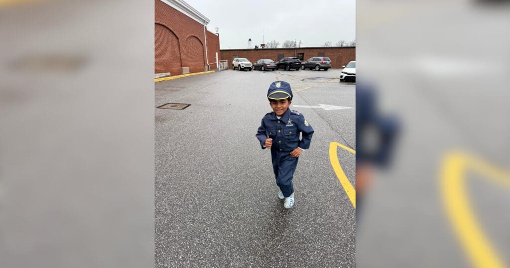 A young child in a police costume runs joyfully in a parking lot. The overcast sky and brick building background suggest a playful, cloudy day scene.