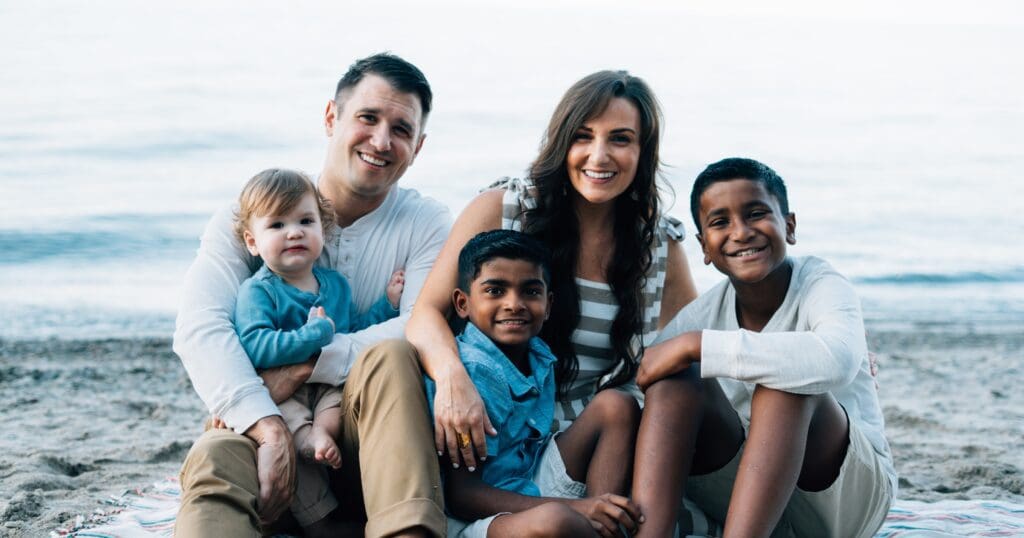 A family sits on a beach, smiling warmly. Parents on the edges, with three children between them. Ocean waves gently in the background, creating a serene atmosphere.