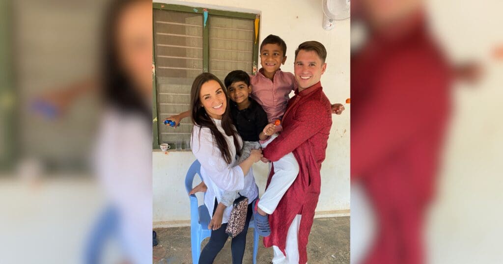 A smiling couple holds two children in an embrace. The scene is joyful, with the adults wearing traditional outfits and the children looking happy. They're indoors, beside a window.