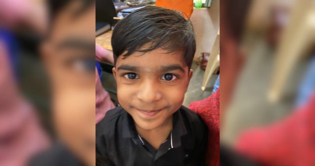 A young child with dark hair and a bright smile looks directly at the camera, wearing a black shirt. The setting is cozy and warm, evoking a cheerful mood.