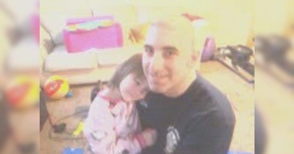 A man and young girl are smiling indoors. The girl, in pajamas, leans affectionately on the man's shoulder. Toys and a sofa are visible in the blurred background.