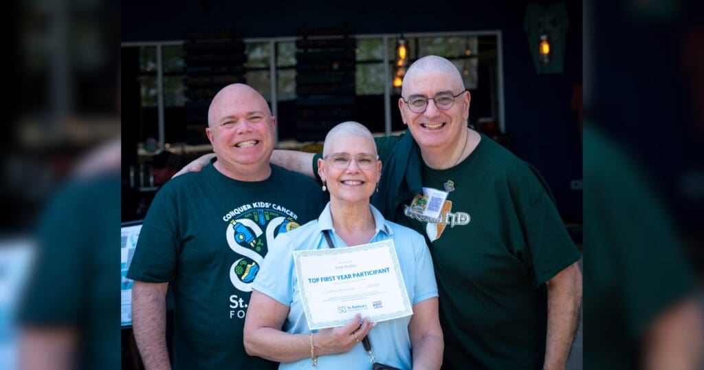 Three smiling people with shaved heads are posing together outdoors. The person in the center holds a certificate labeled "Top First Year Participant."