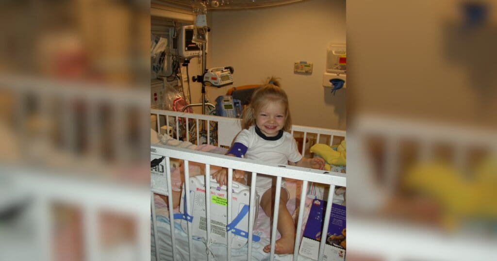 A young child with blonde hair sits smiling in a hospital crib. Medical monitors and equipment are visible in the background, suggesting a hospital setting.