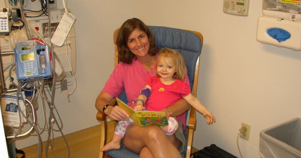 A woman and a young child sitting in a hospital room, smiling. The woman reads a colorful book to the child, who has bandages on her arm. Cozy and caring atmosphere.