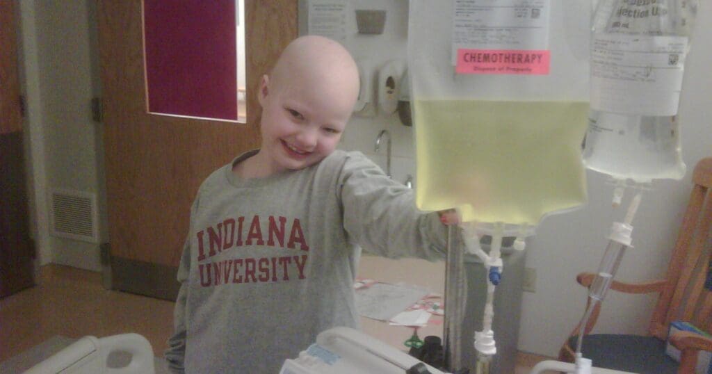 A smiling child in a hospital room, wearing an "Indiana University" shirt, stands beside a chemotherapy IV drip, conveying hope and resilience.