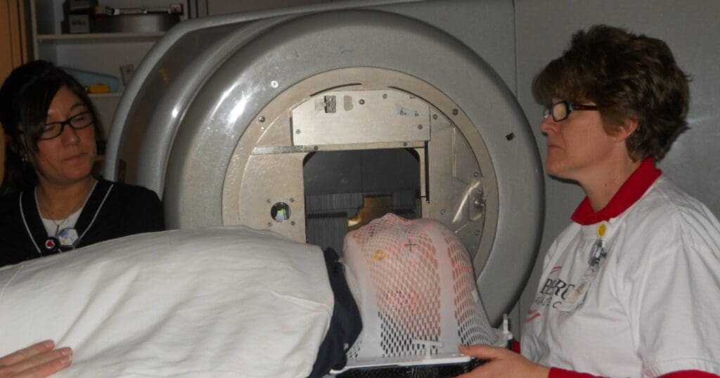 Two medical professionals assist a patient in a CT scanner. The patient wears a mesh mask, conveying a sense of care and focus on the procedure.