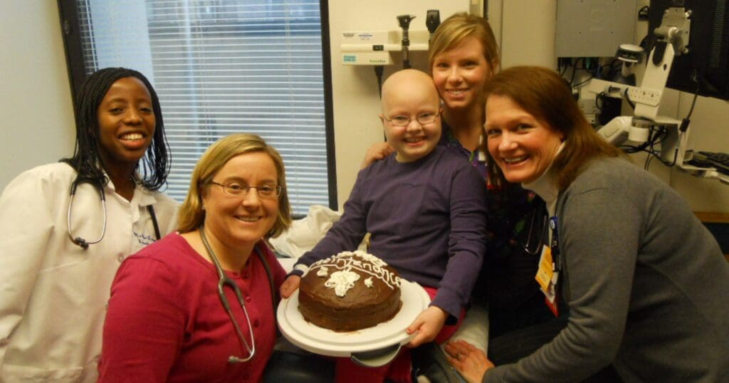 A child in a hospital room, holding a chocolate cake, is surrounded by four smiling women, one in a lab coat, conveying warmth and celebration.