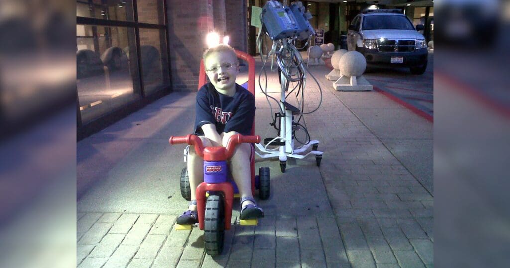 A child with glasses joyfully rides a red tricycle outside on a brick path, with an IV stand beside them. The scene is well-lit and cheerful.