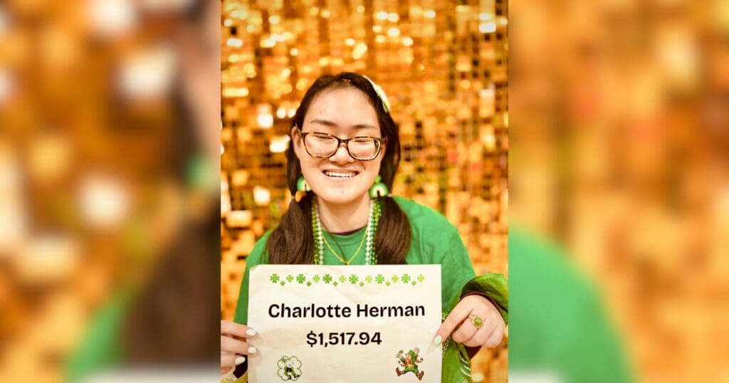 A woman in a green outfit holds a sign displaying the name "Charlotte Hermann $1,517.94."