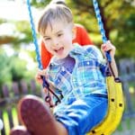 A smiling young boy in a blue plaid shirt swings outdoors, gripping blue ropes as his feet reach toward the camera; bright sunlight and greenery create a joyful, playful mood.